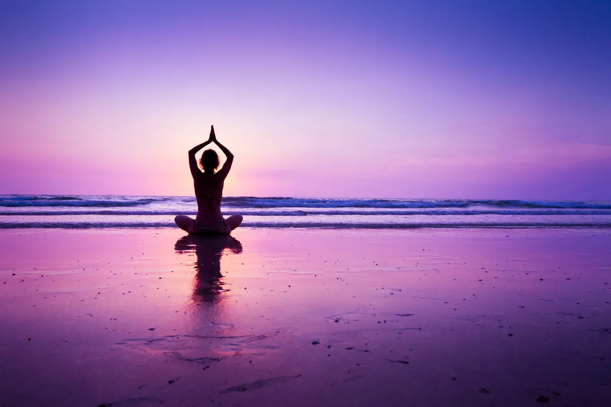A woman sits cross-legged in meditation on wet sand at the ocean's edge, silhouetted against a violet sunset.