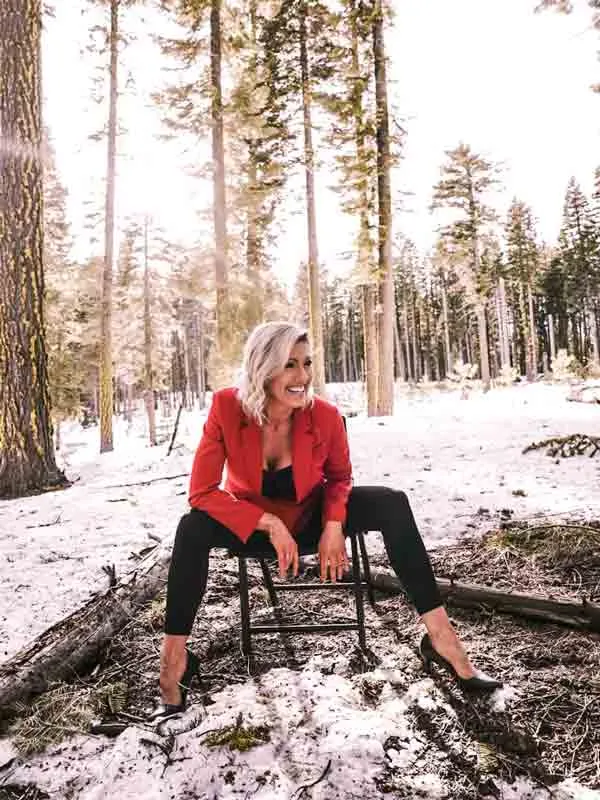 Joy August sits on a stool in a sunlit, snow-dusted pine forest, wearing a red blazer and laughing off to one side.